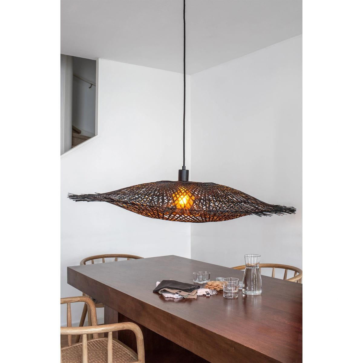 Lampa wisząca Kumo w kolorze czarnym z kloszem o organicznym kształcie, wykonanym z plecionki. Zawieszona na czarnym kablu, wisi nad drewnianym stołem, emitując ciepłe światło. Idealna do nowoczesnych wnętrz.