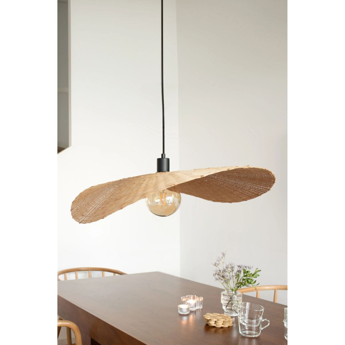Lampa wisząca Rayo z kloszem w kształcie dwóch fal, wykonana z naturalnego materiału, zawieszona na czarnym przewodzie. Widoczna żarówka E27, lampa umieszczona nad drewnianym stołem, w otoczeniu szklanych naczyń i świec.