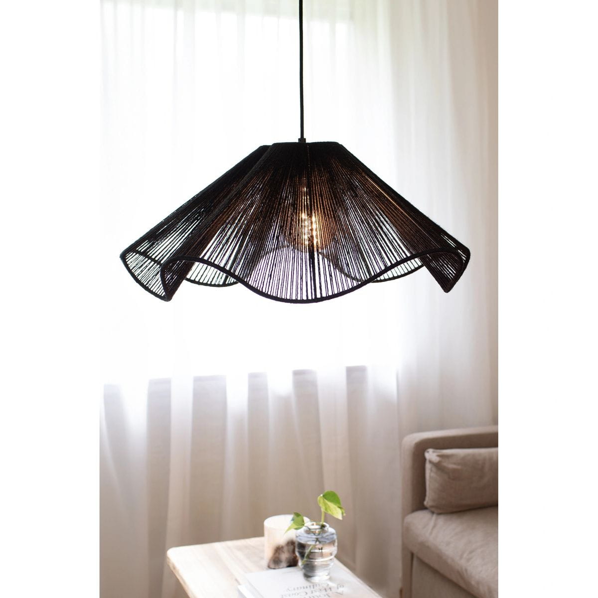 Lampa wisząca Nami w kolorze czarnym z kloszem o falowanym kształcie, wykonana z cienkich przędzy. Widoczna żarówka E27 w centralnej części. Zawieszona nad stolikiem w jasnym wnętrzu z zasłonami w tle.