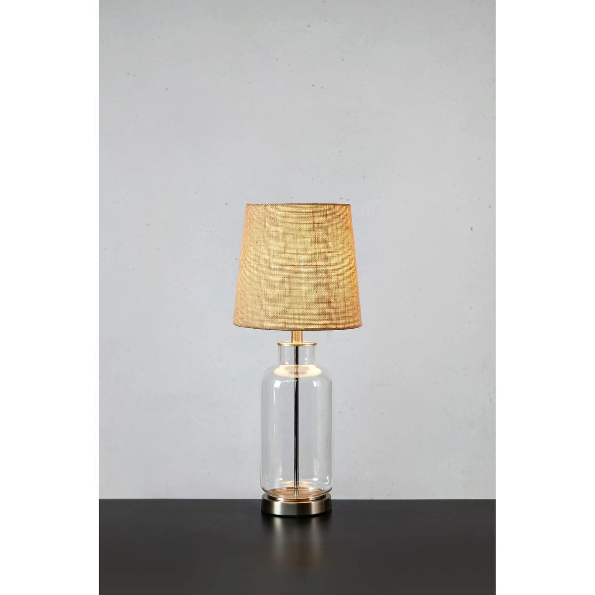 Lampa stołowa Costero z kloszem w kolorze naturalnym, umieszczona na przezroczystym, szklanym korpusie. Ujęcie frontalne, lampa stoi na czarnym blacie. Widoczne detale, w tym metalowa podstawa i elegancki kształt klosza.