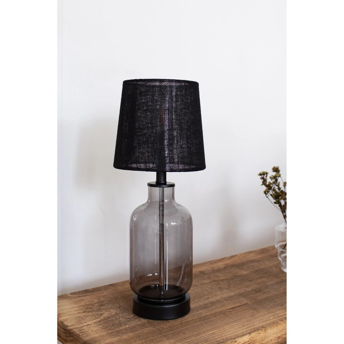 Lampa stołowa Costero z dymną szklaną podstawą i czarnym kloszem z tkaniny. Ujęcie z boku, stojąca na drewnianym stole. Wysoka, o cylindrycznym kształcie, z metalowym stelażem. Tło jasne, minimalistyczne.
