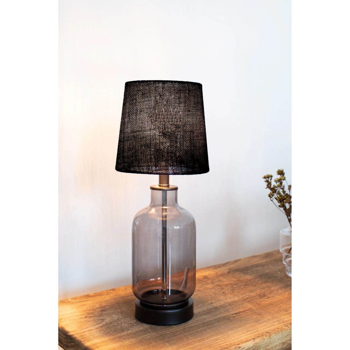 Lampa stołowa Costero z dymnym, szklanym kloszem i czarnym abażurem. Ustawiona na drewnianym stole, o cylindrycznym kształcie z metalowym elementem w kolorze złotym. Wysokość lampy optymalna do użytku domowego.