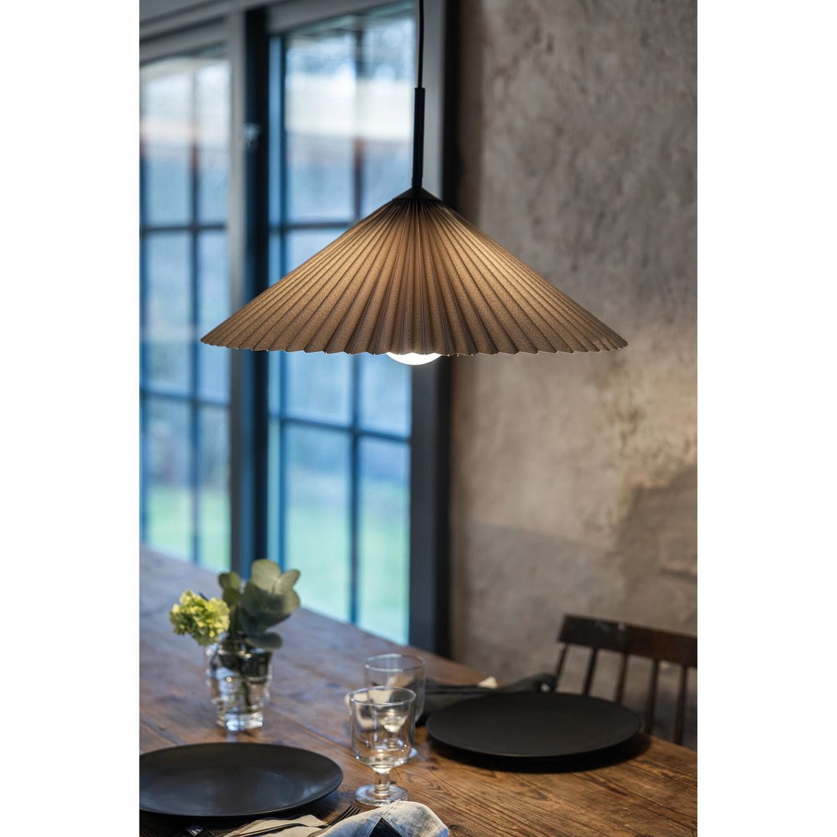 Lampa wisząca Plisado w kolorze czarnym i szarym, z kloszem w kształcie stożka z fałdami. Zawieszona nad drewnianym stołem, oświetla stół z czarnymi talerzami, szklankami i rośliną w wazonie. Kąt ujęcia z boku, ukazujący detale klosza i otoczenie.