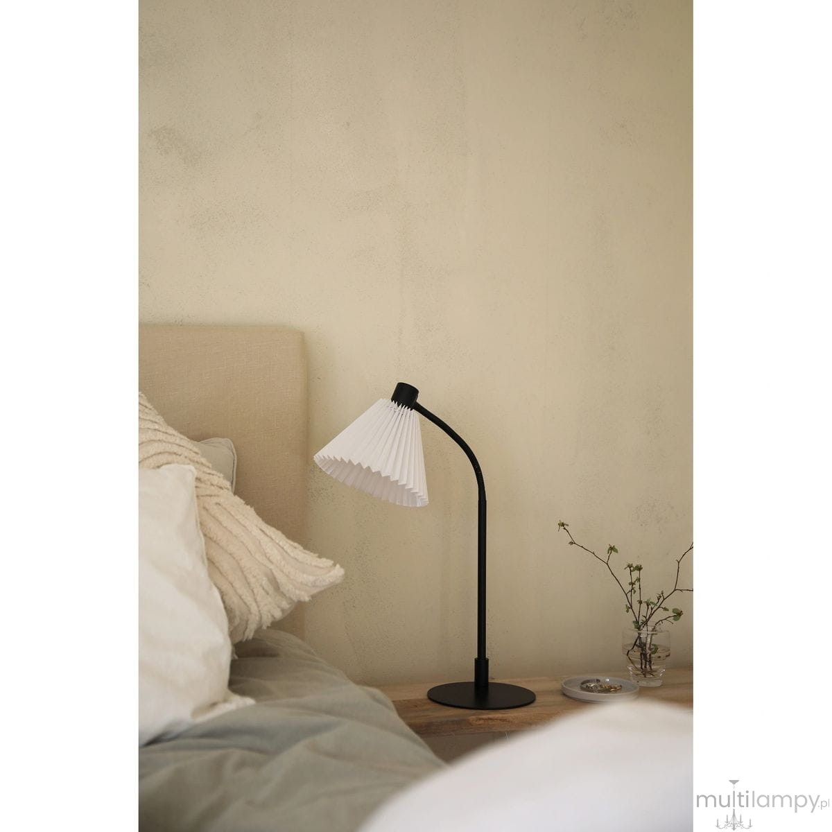 Lampa stołowa Mira z kloszem w białym kolorze i czarną podstawą, ustawiona na drewnianym stoliku obok łóżka. Klosz ma teksturowaną, fałdowaną formę. W tle widoczna beżowa ściana oraz poduszki na łóżku.