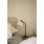 Lampa stołowa Mira z kloszem w białym kolorze i czarną podstawą, ustawiona na drewnianym stoliku obok łóżka. Klosz ma teksturowaną, fałdowaną formę. W tle widoczna beżowa ściana oraz poduszki na łóżku.