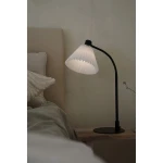Lampa stołowa Mira w kolorze czarnym i białym z kloszem w kształcie stożka, wykonanym z plisowanego materiału. Ustawiona na drewnianym stoliku obok łóżka, z elastycznym ramieniem umożliwiającym regulację kąta.