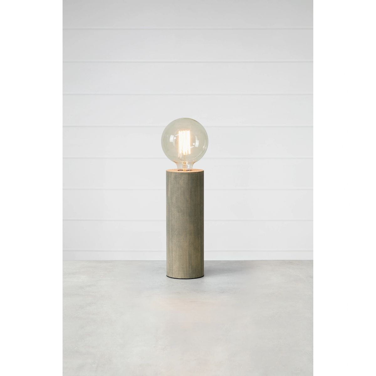 Lampa stołowa Legna z szarym, cylindrycznym słupkiem z betonu i dużą, okrągłą żarówką E27. Ujęcie frontalne, lampa stoi na płaskiej powierzchni, w tle jasne, jednolite ściany.