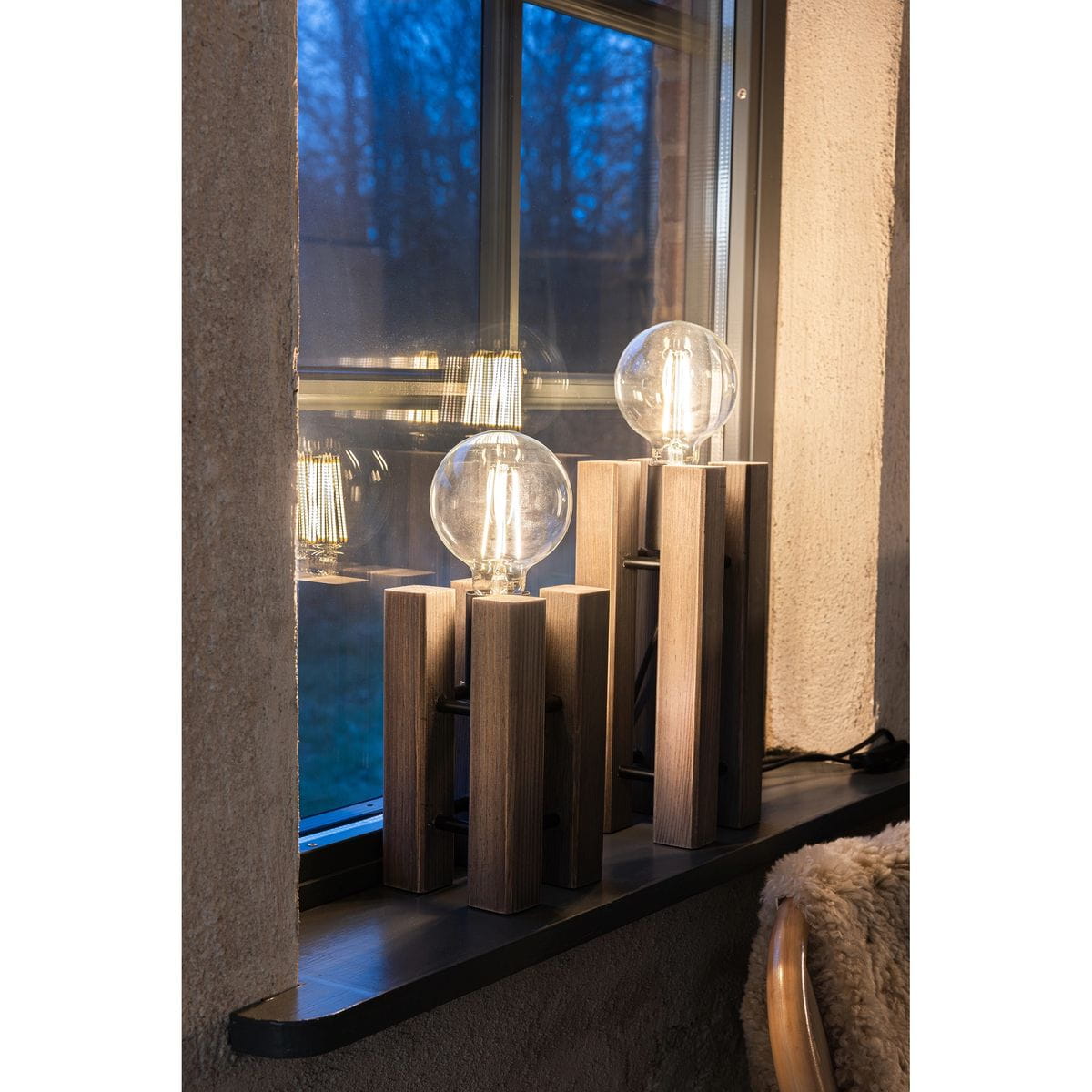 Lodge lampa stołowa 1xE27 brązowa, z drewnianym stelażem w formie prostokątnych słupków. Na każdym słupku znajduje się okrągła, przezroczysta żarówka. Lampa ustawiona na parapecie, w tle widoczna niebieska przestrzeń przez okno.