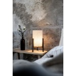 Lampa stołowa Eruca w kolorze brązowym i beżowym z abażurem z materiału, umieszczona na drewnianym stoliku. Obok znajduje się ciemny wazon z białą różą. Tło w odcieniach szarości z widoczną ceglaną ścianą.