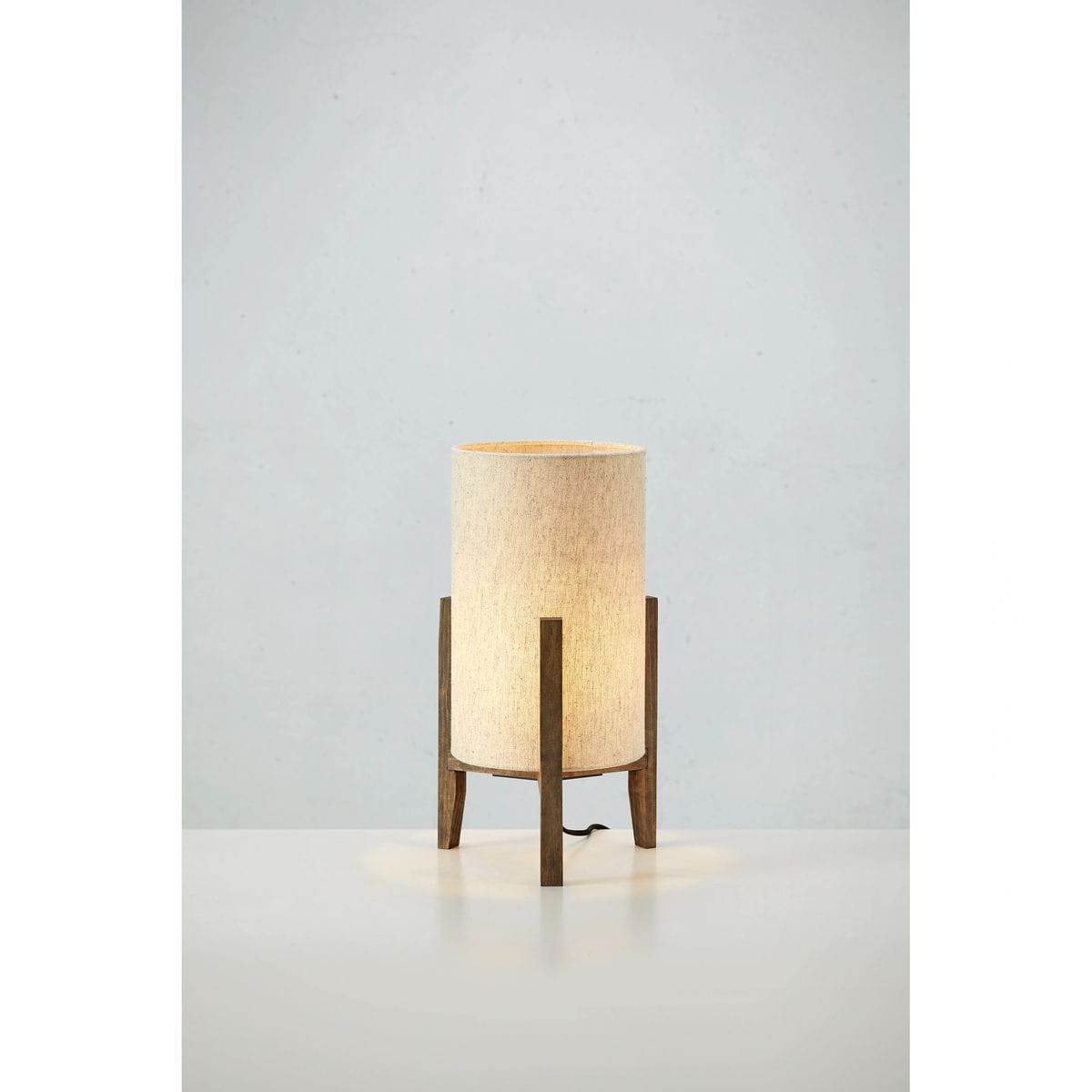 Lampa stołowa Eruca z brązowym drewnianym stelażem i beżowym, tekstylnym kloszem. Ustawiona na płaskiej powierzchni, o cylindrycznym kształcie, z widocznym przewodem zasilającym. Świeci ciepłym światłem, tworząc przytulną atmosferę.
