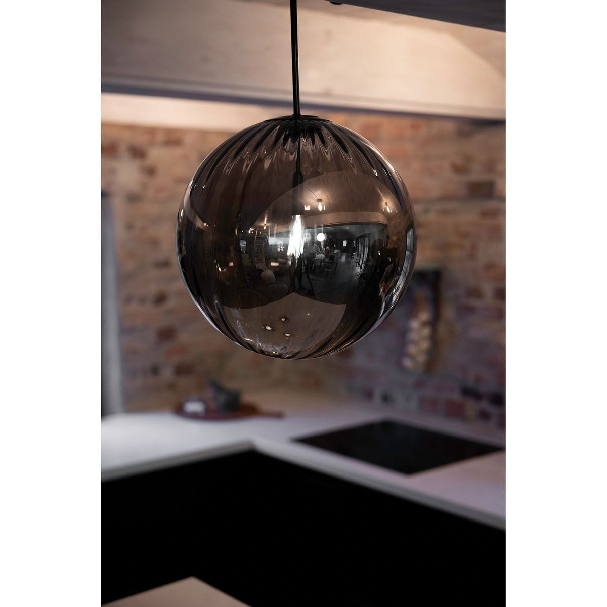 Czarna lampa wisząca Fengari z dymionym kloszem, w kształcie kulistym, zawieszona na cienkim przewodzie. Widoczny połysk szkła oraz subtelne refleksy. Tło to nowoczesna kuchnia z jasnym blatem i ciemną zabudową.