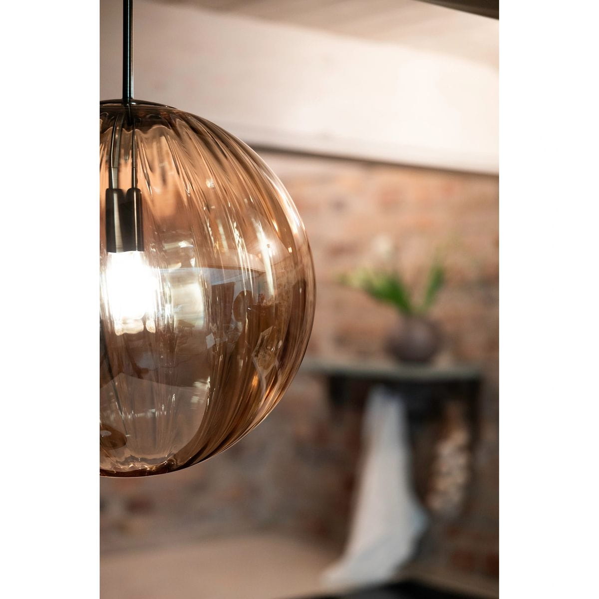 Lampa wisząca Fengari 1xE14 w kolorze bursztynowym, z antycznym mosiądzu. Okrągła, szklana kula z wyraźnym wzorem, zawieszona na czarnym przewodzie. W tle widoczna ceglana ściana oraz dekoracyjny element z roślinami.