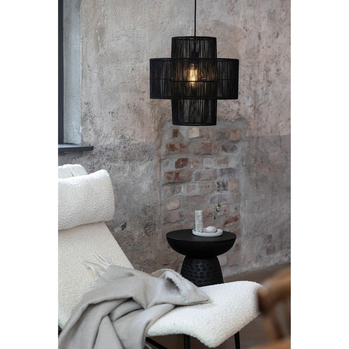 Lampa wisząca Soga w kolorze czarnym, z miejscem na żarówkę E27, o nowoczesnym, ażurowym kształcie. Zawieszona nad czarnym stolikiem, w otoczeniu jasnej leżanki i dekoracji, na tle surowej, ceglanej ściany.