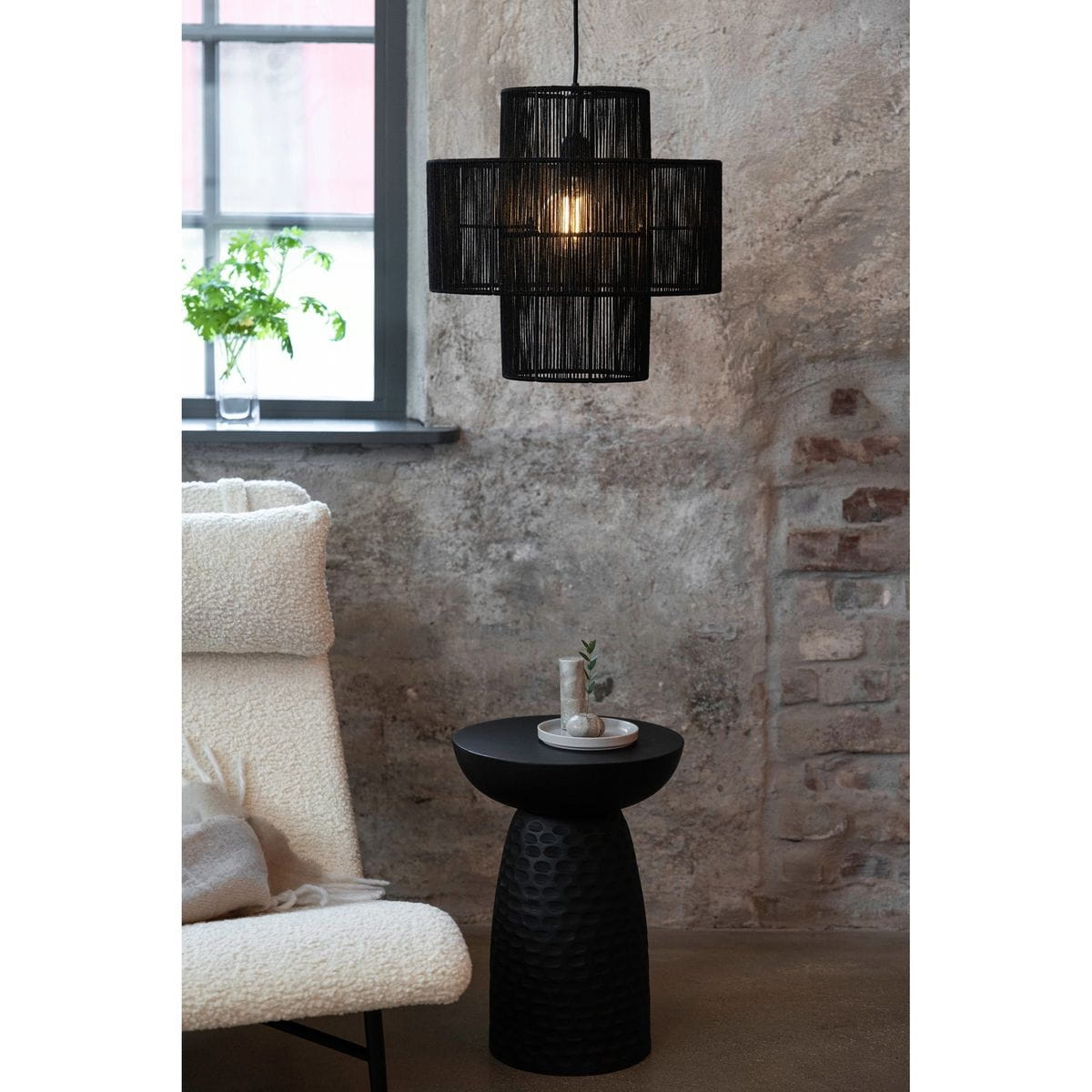 Lampa wisząca Soga w kolorze czarnym z jednym gniazdem E27, o strukturalnej formie z plecionki. Zawieszona nad okrągłym stolikiem o fakturze przypominającej plastry miodu, w aranżacji z jasnym fotelem i rośliną w tle.