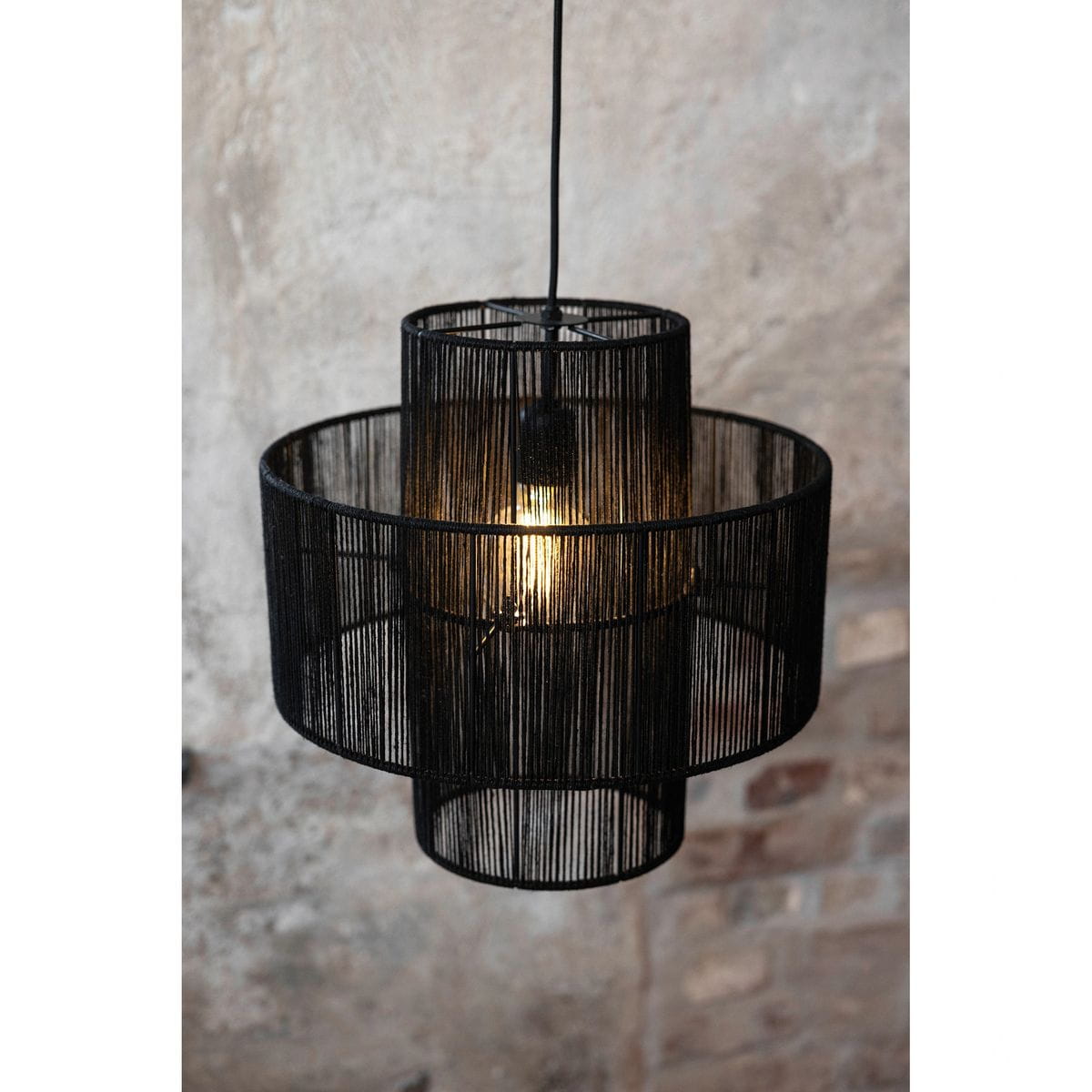 Lampa wisząca Soga 1xE27 w kolorze czarnym z metalową konstrukcją, składająca się z dwóch okrągłych kloszy z cienkimi, pionowymi linkami. Źródło światła umieszczone wewnątrz, w tle widoczna surowa ściana z cegły.