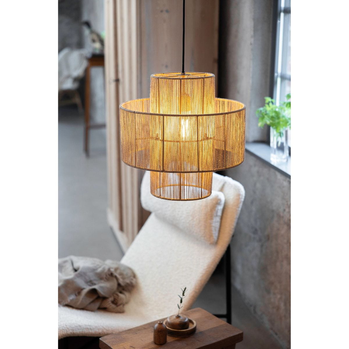Lampa wisząca Soga z jedną oprawą E27, w naturalnym kolorze, z plecioną konstrukcją z cienkich prętów. Zawieszona nad wygodnym fotelem, emituje ciepłe światło. Widoczna w aranżacji z drewnianym stolikiem i roślinnością w tle.