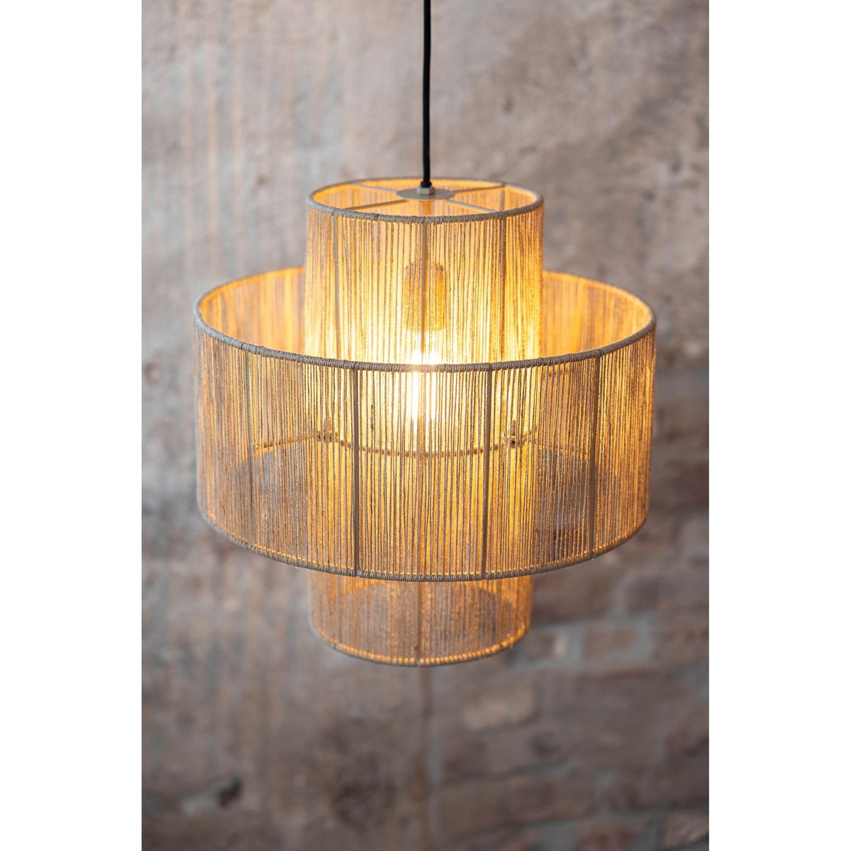 Lampa wisząca Soga z oprawą na jedną żarówkę E27, wykonana z cienkiego, naturalnego materiału, w kształcie dwóch nachodzących na siebie okręgów. Zawieszona na czarnym przewodzie, emitująca ciepłe światło. Tło w odcieniach szarości.