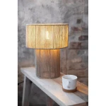 Lampa stołowa Soga z kloszem z naturalnych włókien, umieszczona na okrągłej podstawie. W tle widoczna drewniana półka z białą filiżanką na podkładce. Oświetlenie włączone, tworzy ciepłą atmosferę. Tło z fakturą cegły.
