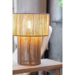 Lampa stołowa Soga z kloszem w naturalnym kolorze, wykonana z materiału z teksturą, z żarówką E27. Ujęcie frontalne, lampa stoi na drewnianym stole, emitując ciepłe światło, otoczona minimalistycznym wnętrzem.