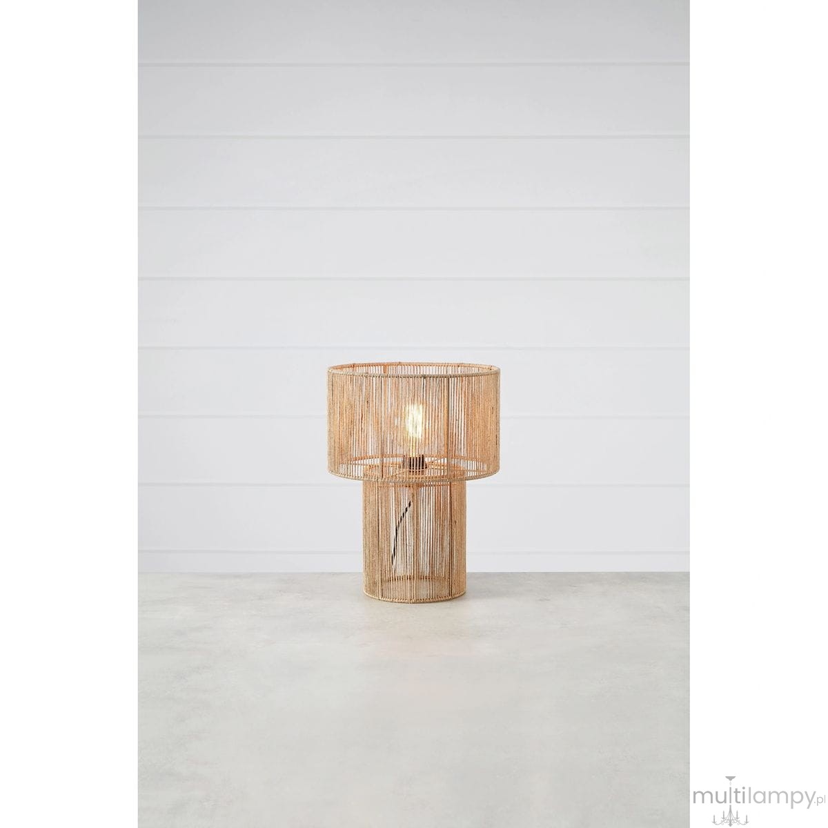 Lampa stołowa Soga z naturalnego materiału, okrągła podsufitka i cylindryczna podstawa. W zestawie żarówka E27. Ujęcie frontalne na szarym tle, lampa świeci, odsłaniając delikatny wzór. Widoczny przewód z włącznikiem.