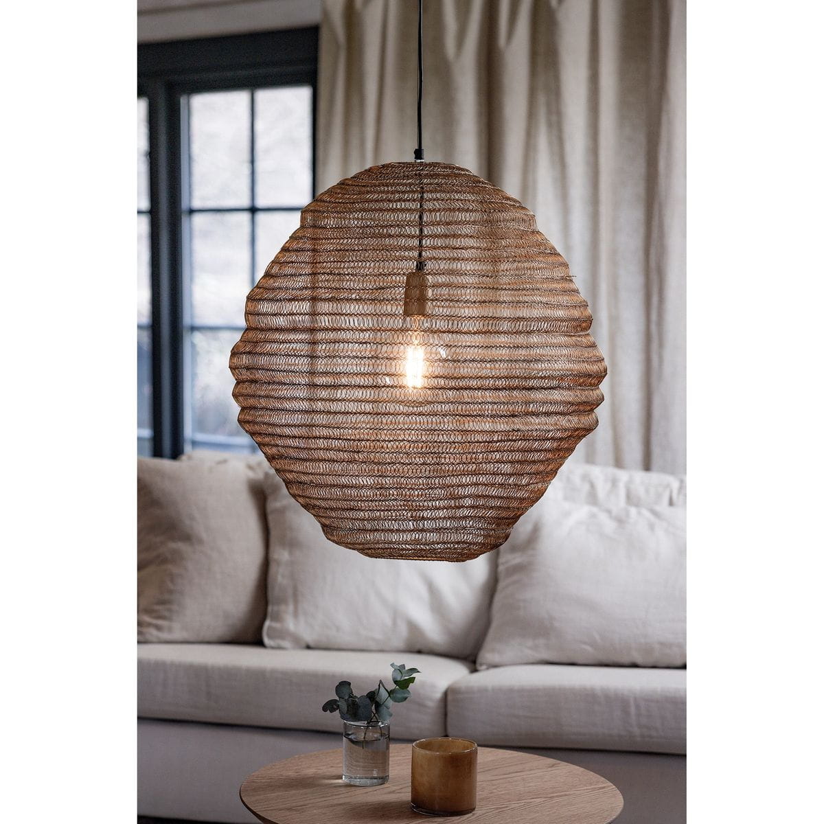 Lampa wisząca Tazza w kolorze czarnym i złotym z kloszem w kształcie okręgu, wykonanym z plecionki. Zawieszona na czarnym przewodzie nad jasną sofą, z widoczną żarówką E27. Tło z zasłonami i oknami, minimalistyczne otoczenie.