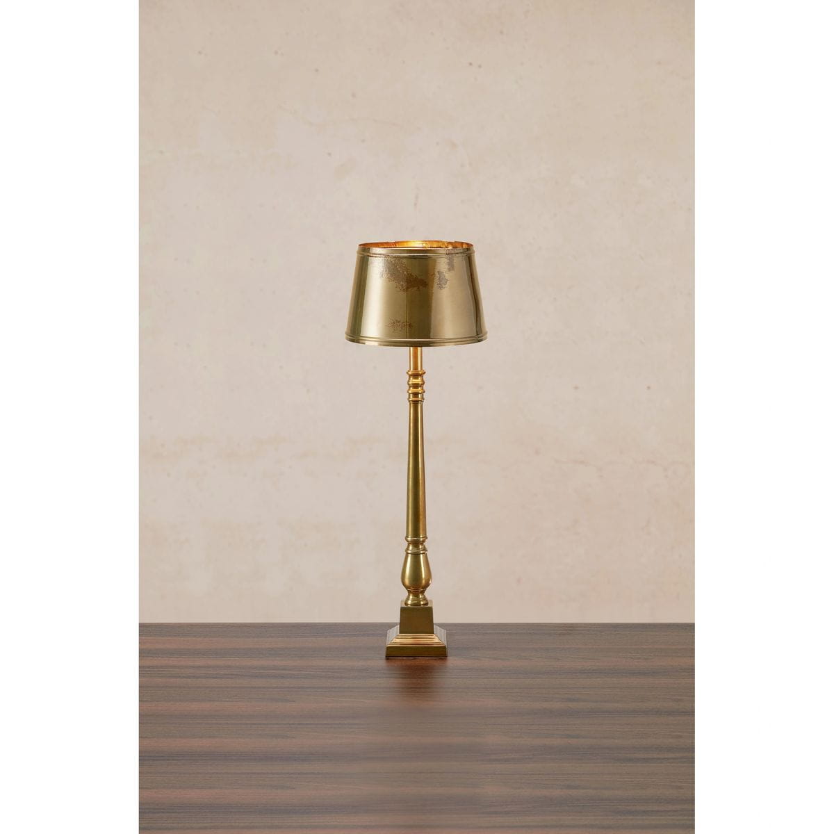 Lampka stołowa Metallo 1xE27 w kolorze antycznego mosiądzu, z cylindrycznym kloszem i smukłą podstawą. Ujęcie frontalne, produkt na drewnianym blacie, z dekoracyjnymi detalami na nodze lampy.