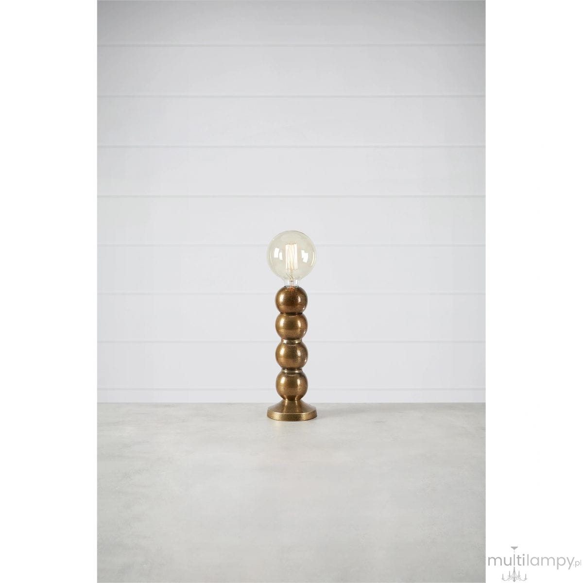 Lampa stołowa Gong w kolorze złotym, z okrągłą podstawą i trzema kulistymi elementami. U góry znajduje się przezroczysta żarówka. Produkt stoi na szarej powierzchni, na tle jasnej ściany.