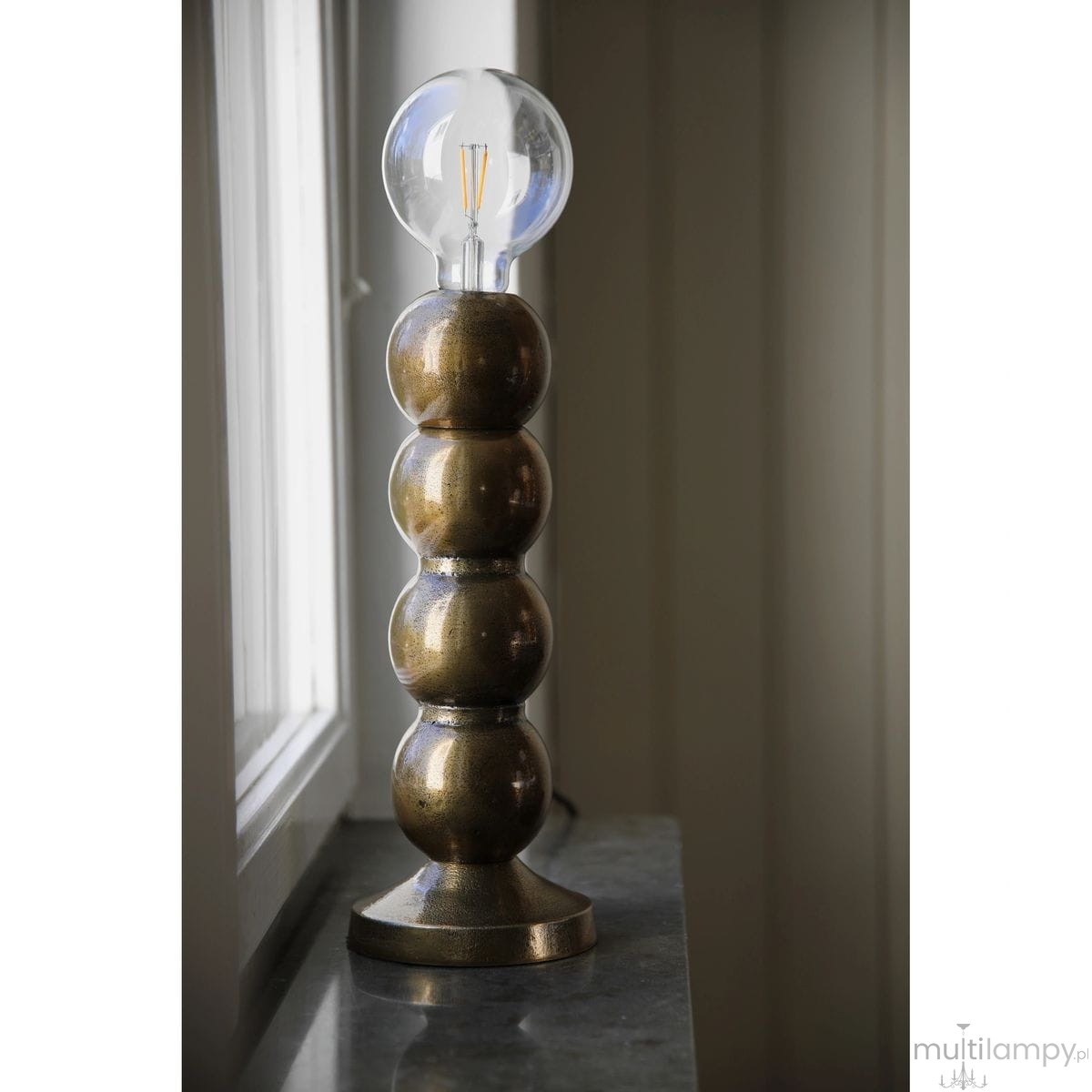 Lampa stołowa Gong 1xE27 w złotym kolorze, o podstawie z okrągłymi, metalicznymi elementami. Przezroczysta żarówka kulista na górze. Produkt ustawiony na kamiennej powierzchni, w tle okno.