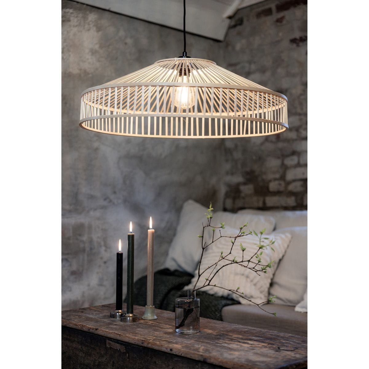 Lampa wisząca Tapa 1xE27 w naturalnym kolorze, o okrągłym kształcie z plecionki, zawieszona na czarnym kablu. W tle widoczne świece i gałązka w szklanym wazonie na drewnianym stole. Ceglana ściana tworzy rustykalne tło.