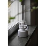 Lampa stołowa Kandella z białą podstawą i przezroczystym kloszem. Kształt cylindryczny, z dekoracyjnym białym detalem u podstawy. Umieszczona na ciemnym blacie obok okna, z widoczną żarówką E14 wewnątrz.