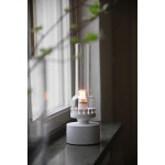 Lampa stołowa Kandella w białym kolorze z przeźroczystym kloszem, z widoczną żarówką E14. Ujęcie z boku, stojąca na oknie, z okrągłą bazą i dekoracyjną obręczą. Czyste tło, naturalne światło, dodające przytulności.