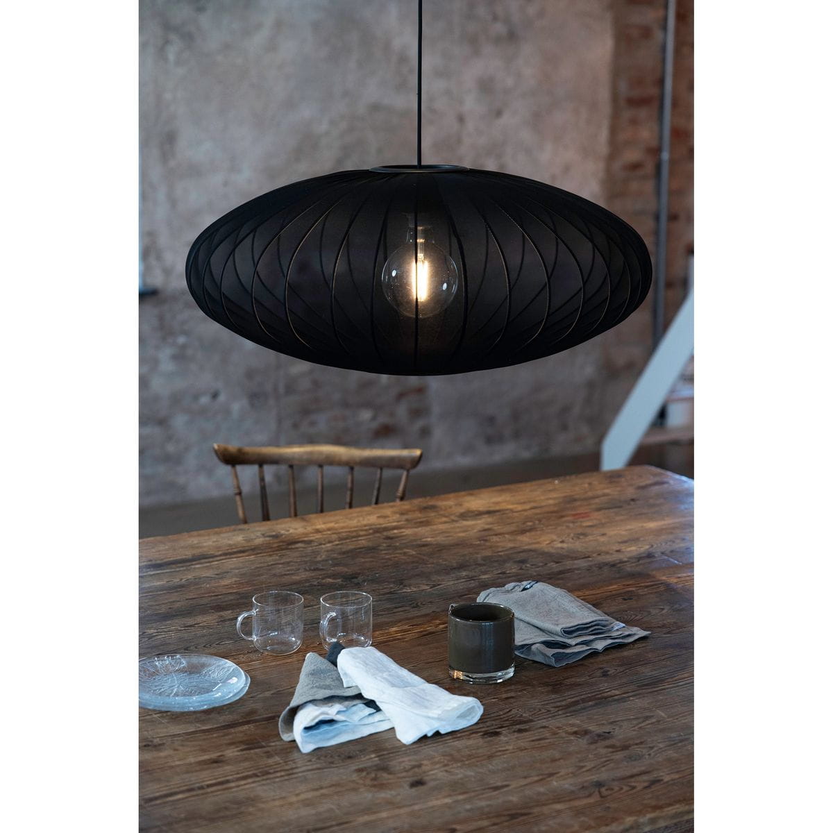 Czarna lampa wisząca Florence D75 z kloszem w kształcie owalnym, wykonana z cienkiego materiału, z widoczną żarówką E27. Powieszona nad drewnianym stołem, w otoczeniu szklanych kubków i serwetek, na tle surowej ściany.
