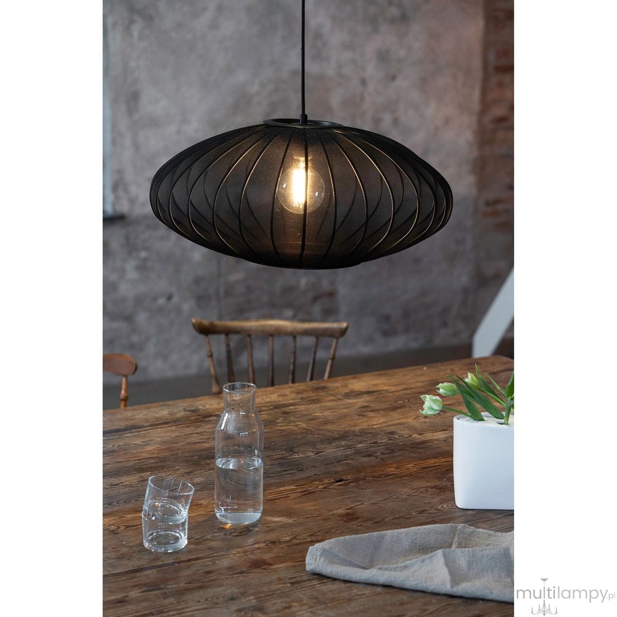 Czarna lampa wisząca Florence D60 z kloszem w formie ażurowej kopuły, zawieszona nad drewnianym stołem. W środku widoczna żarówka E27. W tle szklany dzbanek i filiżanka, na stole również widać biały doniczkę z roślinami.