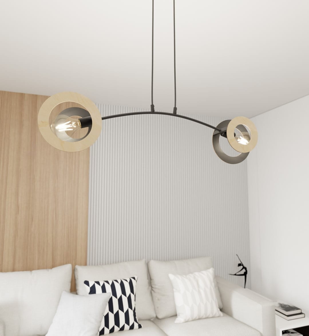 Lampa wisząca Skiva z dwoma okrągłymi kloszami, czarna metalowa konstrukcja z jasnym drewnem. Ujęcie frontalne, zawieszona na cienkich linkach. Tło w jasnych odcieniach, w aranżacji salonu z jasną sofą i poduszkami.