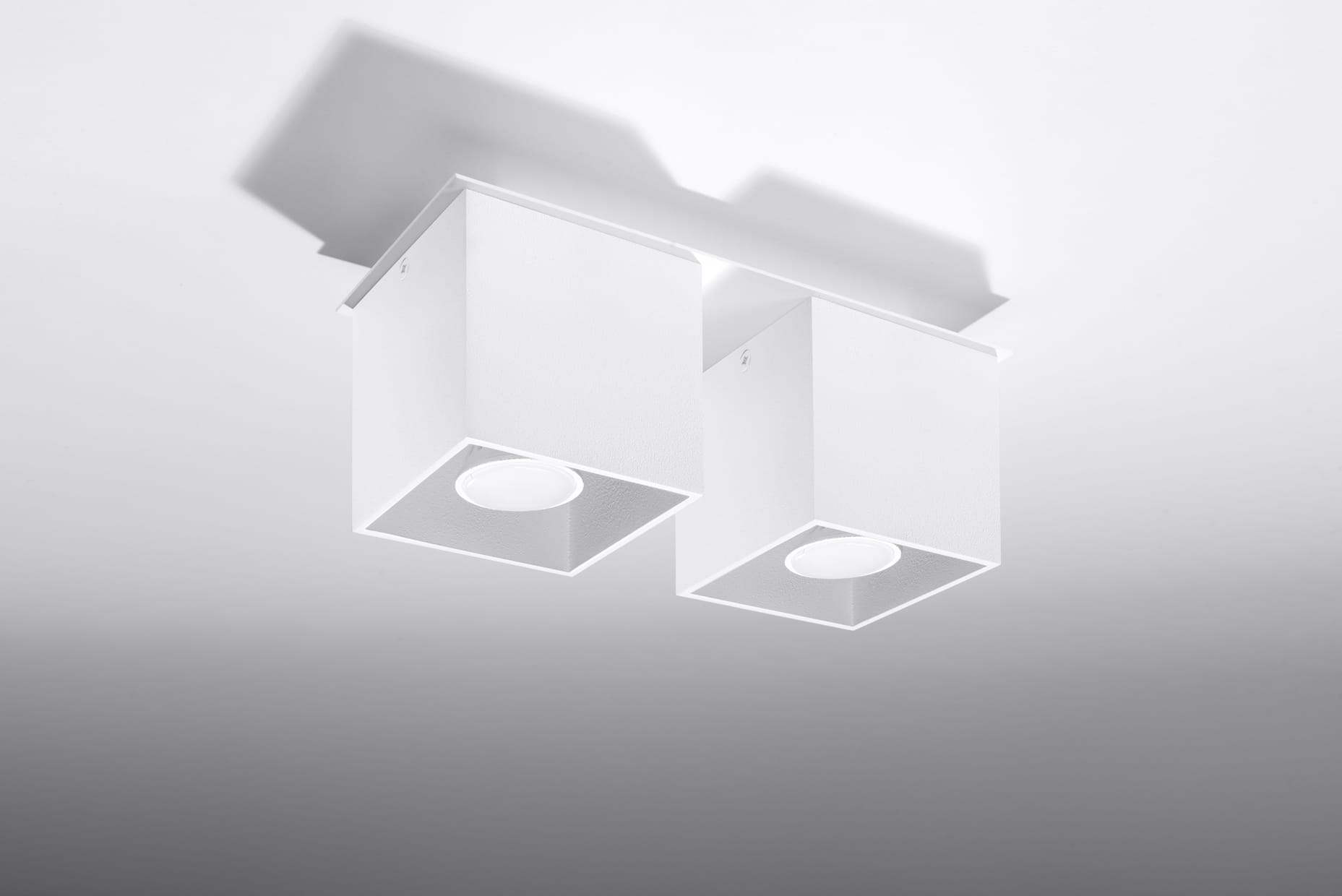 Lampa sufitowa 2-punktowa w kolorze białym, składająca się z dwóch kwadratowych kloszy z osłoną na żarówki. Ujęcie z dołu ukazuje detale konstrukcji i kształt. Produkt montowany do sufitu, z prostym, nowoczesnym designem.
