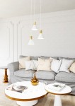 Lampa wisząca Rosa z trzema kloszami w kształcie stożków, w kolorze białym i złotym. Zawieszona na białych linkach, umieszczona nad okrągłym stołem kawowym w nowoczesnym wnętrzu z szarym sofą i dekoracjami.