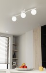 Lampa sufitowa Opus z trzema kulistymi kloszami w kolorze opalowym, zamontowana na białej, zakrzywionej konstrukcji. Ujęcie z dołu, pokazujące lampę w nowoczesnym wnętrzu z widocznymi półkami i dekoracjami.