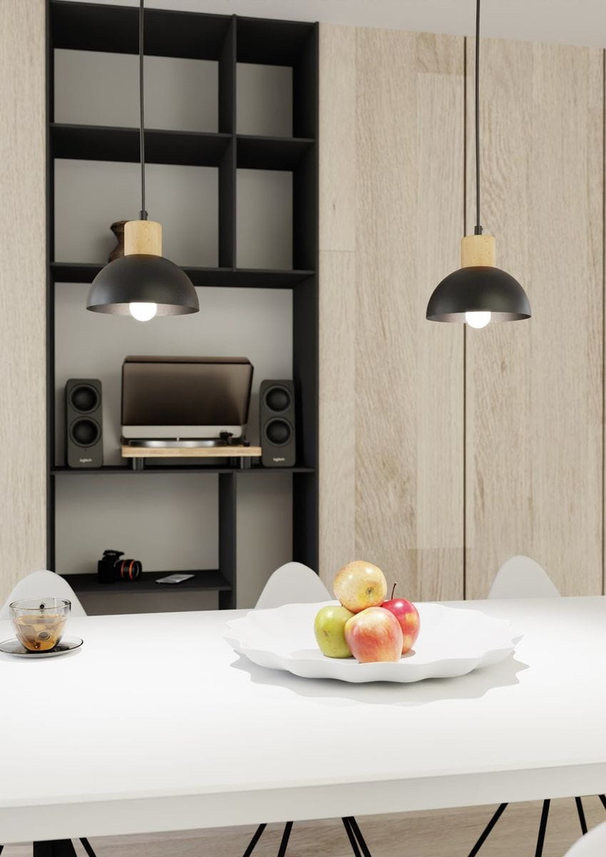 Lampa wisząca Seco 1xE27 w kolorze czarnym z drewnianym elementem, zawieszona na czarnym przewodzie. Ujęcie z boku, w tle widoczna półka z elektroniką. Produkt o zaokrąglonym kształcie, umieszczony nad białym stołem z talerzem z owocami.