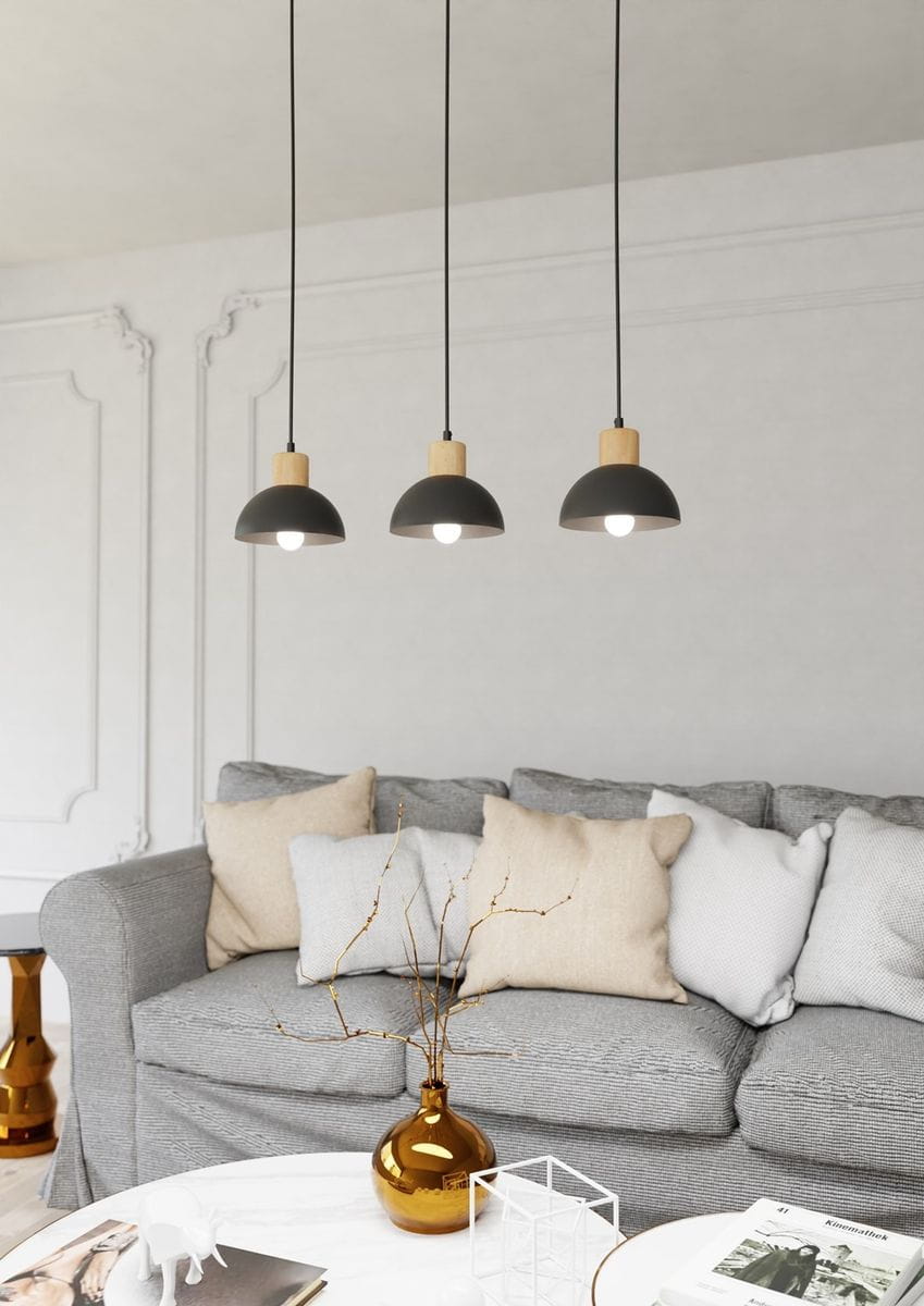 Lampa wisząca Seco z trzema kloszami w kolorze czarnym i jasnym drewnie. Umieszczona pod sufitem, zwisająca na cienkich kablach. W tle widoczna szara kanapa z poduszkami oraz złota dekoracyjna waza na stole.