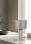 Lampa stołowa Canio z szarym abażurem i naturalnym, teksturowanym stelażem, stojąca na jasnym blacie. Widoczne są kable zasilające oraz minimalistyczne tło z zasłonami i dekoracyjnym naczyniem.