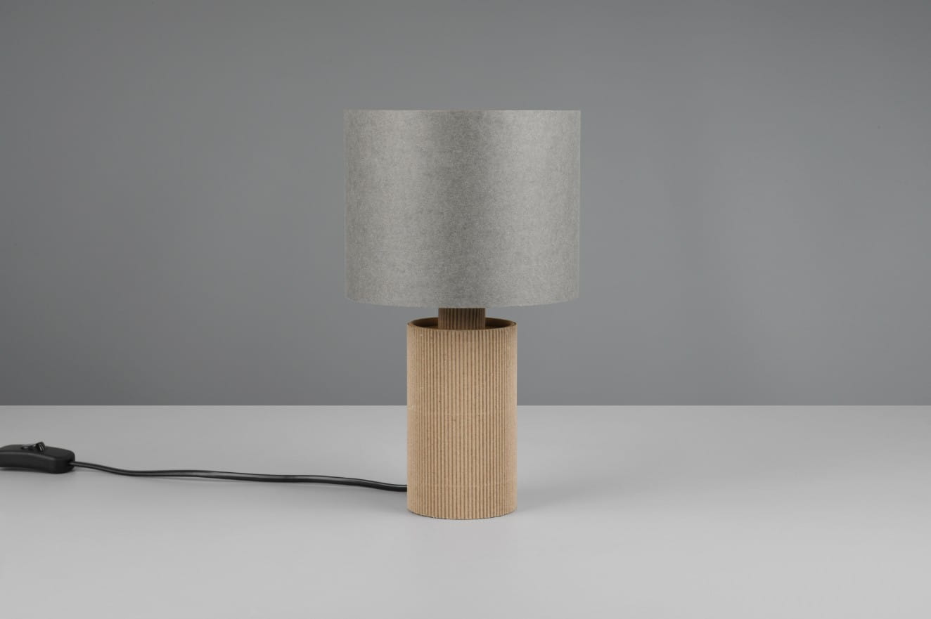 Lampa stołowa Canio z szarym abażurem z materiału i naturalną, teksturowaną podstawą w kształcie cylindra. Produkt stoi na białym blacie, obok widoczny przewód z włącznikiem. Tło w odcieniu szarości.