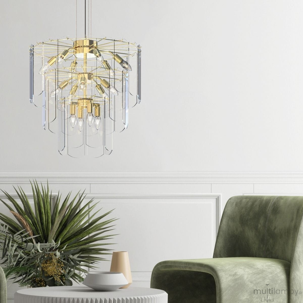 Lampa wisząca Nira w kolorze złotym z 14 miejscami na żarówki E14. Ozdobna konstrukcja z przezroczystymi akrylowymi elementami. Zawieszona w nowoczesnym wnętrzu nad okrągłym stołem, obok zielonej rośliny i sofą.