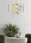 Lampa wisząca Nira 14xE14 w kolorze złotym, z przezroczystymi elementami. Ujęcie frontalne, lampa zawieszona nad okrągłym stołem. W tle rośliny dekoracyjne oraz zielone krzesła. Wysoka jakość wykonania, nowoczesny design.