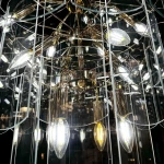 Złota lampa wisząca Nira z 14 miejscami na żarówki E14, o eleganckim, transparentnym kloszu, z widocznymi żarówkami w różnych odcieniach. Kąt ujęcia zbliżeniowy, ukazujący detale konstrukcji i połysk materiału.
