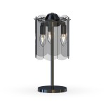 Lampa stołowa Nira w kolorze czarnym z chromowanym wykończeniem, wyposażona w trzy gniazda E14. Posiada przezroczystą osłonę z zaokrąglonymi krawędziami, stojącą na okrągłej podstawie. Ujęcie frontalne, ukazujące całość produktu.