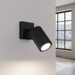 Kinkiet Karbon 1xGU10 w kolorze czarnym, z cylindryczną, żłobioną oprawą lampy, zamontowany na ścianie. Źródło światła skierowane w dół, na tle jasnej ściany i okna w tle.