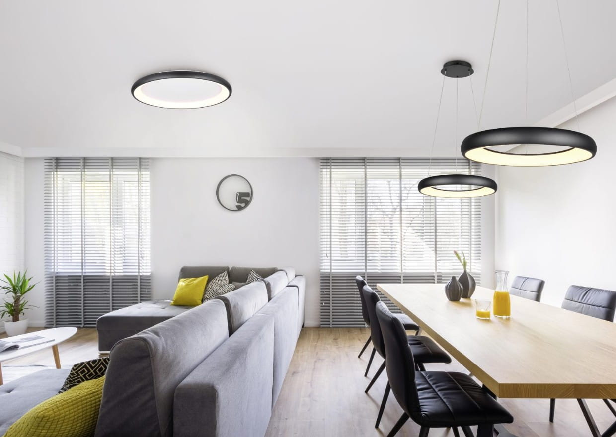Lampa wisząca Cardona w kolorze czarnym, z jedną żarówką LED 38W, o okrągłym kształcie. Zawieszona na cienkich linkach, emituje ciepłe światło. W tle nowoczesne wnętrze z szarym sofą i drewnianym stołem.