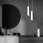 Lampa wisząca Turyn IP44 w kolorze białym, z cylindrycznym kształtem, zawieszona na cienkich przewodach. W tle widoczna okrągła lustro oraz stół z szarym blatem. Na stole brązowe pojemniki oraz wazon z gałązkami.