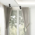 Czarna lampa sufitowa Turyn IP44 z jednym spotem GU10, zamontowana na suficie, skierowana w dół. Tło z zasłonami i oknem, co sugeruje zastosowanie w domowym wnętrzu. Lampa ma minimalistyczny design, idealna do oświetlenia przestrzeni.
