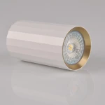 Lampa sufitowa Nicea w kształcie tuby, kolor biały z złotym wykończeniem. Wykonana z matowego materiału, z widocznym źródłem światła GU10. Ujęcie w zbliżeniu, ukazujące detal konstrukcji i reflektor. Tło jednolite, szare.